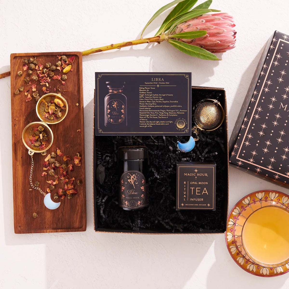 The Libra Astrology Tea Gift Set embraces a tea-themed design and features a &quot;Libra&quot; card, a canister of Moonlight White Tea, a gold infuser, and a floral display. Its box is adorned with starry patterns, complemented by herbs and flowers arranged on a wooden tray for an enchanting presentation.