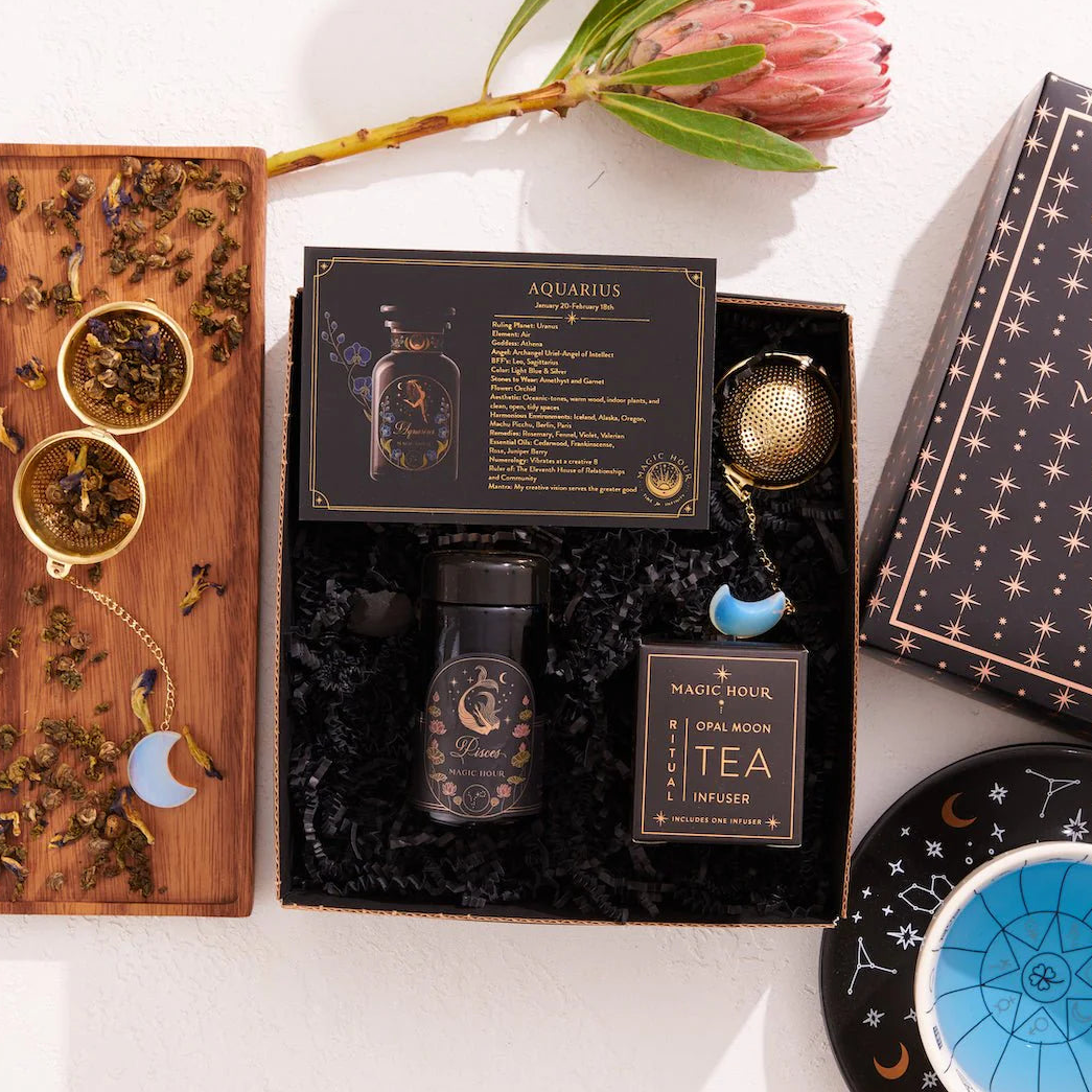 Introducing the Pisces Astrology Tea Gift Set: a celestial collection for tea lovers. It includes a bottle of Pisces tea, Magic Hour infuser, loose tea on a wooden board with opalite moon tea strainer, celestial-themed box, and a black dish adorned with stars. A pink protea flower completes this enchanting ensemble.