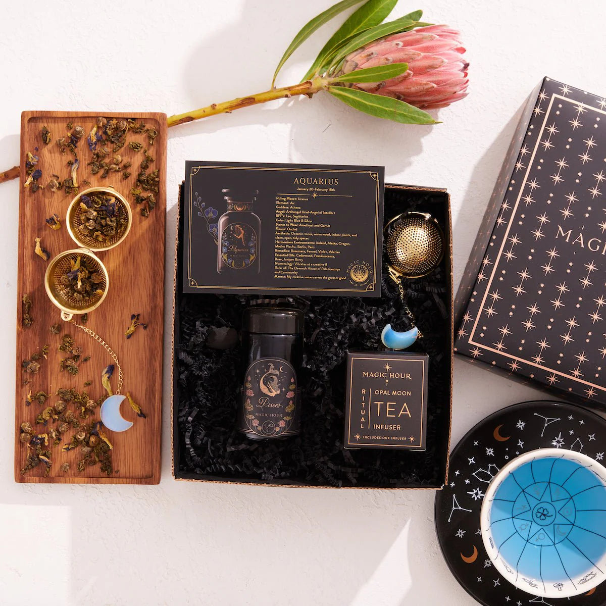 Introducing the Pisces Astrology Tea Gift Set: a celestial collection for tea lovers. It includes a bottle of Pisces tea, Magic Hour infuser, loose tea on a wooden board with opalite moon tea strainer, celestial-themed box, and a black dish adorned with stars. A pink protea flower completes this enchanting ensemble.