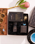 Introducing the Pisces Astrology Tea Gift Set: a celestial collection for tea lovers. It includes a bottle of Pisces tea, Magic Hour infuser, loose tea on a wooden board with opalite moon tea strainer, celestial-themed box, and a black dish adorned with stars. A pink protea flower completes this enchanting ensemble.