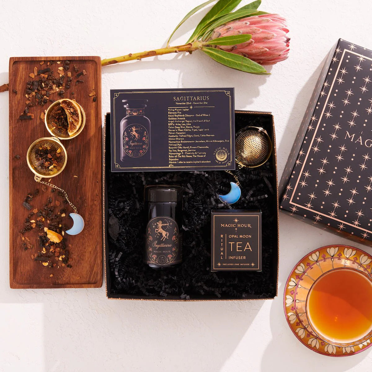 Flat lay of a Sagittarius Astrology Tea Gift Set, which includes a themed card, a jar of loose tea, and a box labeled "Magic Hour Opal Moon Tea & Infuser." A flower and cup of tea rest on a saucer. A wooden tray displays tea leaves alongside an Opalite Moon Tea Strainer adorned with a blue crescent moon charm.