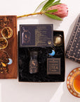 Flat lay of a Sagittarius Astrology Tea Gift Set, which includes a themed card, a jar of loose tea, and a box labeled "Magic Hour Opal Moon Tea & Infuser." A flower and cup of tea rest on a saucer. A wooden tray displays tea leaves alongside an Opalite Moon Tea Strainer adorned with a blue crescent moon charm.