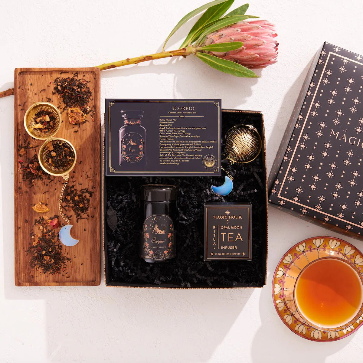 Explore the Scorpio Astrology Tea Gift Set, a heavenly collection that includes a Scorpio-themed tea package with a jar, tea infuser, insightful card, and a magical box on a wooden tray. This set is complemented by a protea flower, two golden spoons filled with loose tea, and a cup nestled in an elegant saucer.