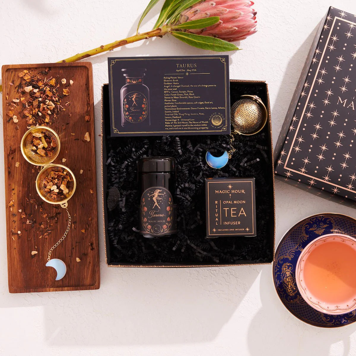 A flat lay of the Taurus Astrology Tea Gift Set, showcasing a sensual and relaxing tea infuser, luxury loose leaf tea, an essential oil bottle, and a zodiac information card. Next to it is a cup with pink tea on a blue saucer and a protea flower as an elegant touch.