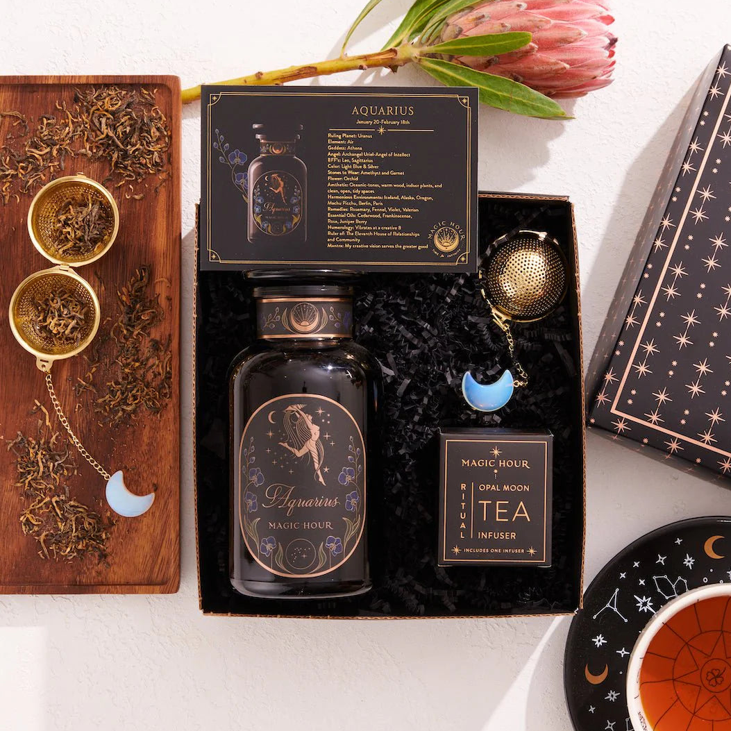 The Aquarius Astrology Tea Gift Set is ideal for astrology enthusiasts. It includes a beautifully designed jar labeled "Aquarius," two opalite moon tea strainers, and a black box embellished with astrological details. The set features a teacup paired with a saucer decorated in a zodiac pattern, and it is complemented by the natural beauty of a protea flower.