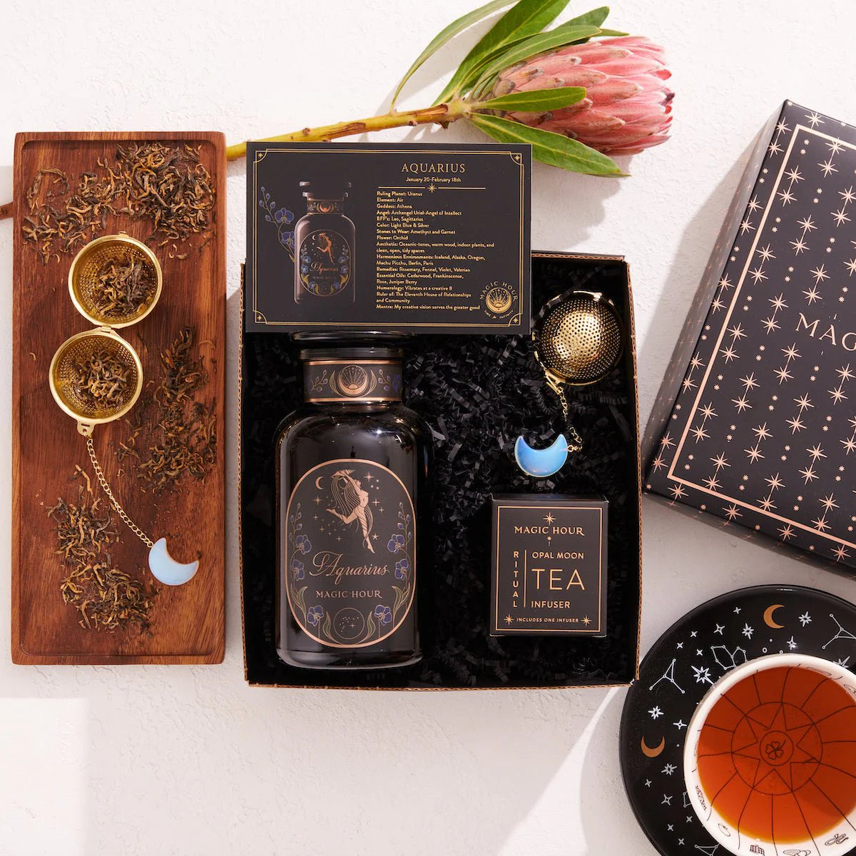 The Aquarius Astrology Tea Gift Set is ideal for astrology enthusiasts. It includes a beautifully designed jar labeled &quot;Aquarius,&quot; two opalite moon tea strainers, and a black box embellished with astrological details. The set features a teacup paired with a saucer decorated in a zodiac pattern, and it is complemented by the natural beauty of a protea flower.