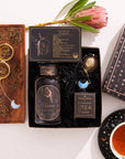 The Aquarius Astrology Tea Gift Set is ideal for astrology enthusiasts. It includes a beautifully designed jar labeled "Aquarius," two opalite moon tea strainers, and a black box embellished with astrological details. The set features a teacup paired with a saucer decorated in a zodiac pattern, and it is complemented by the natural beauty of a protea flower.