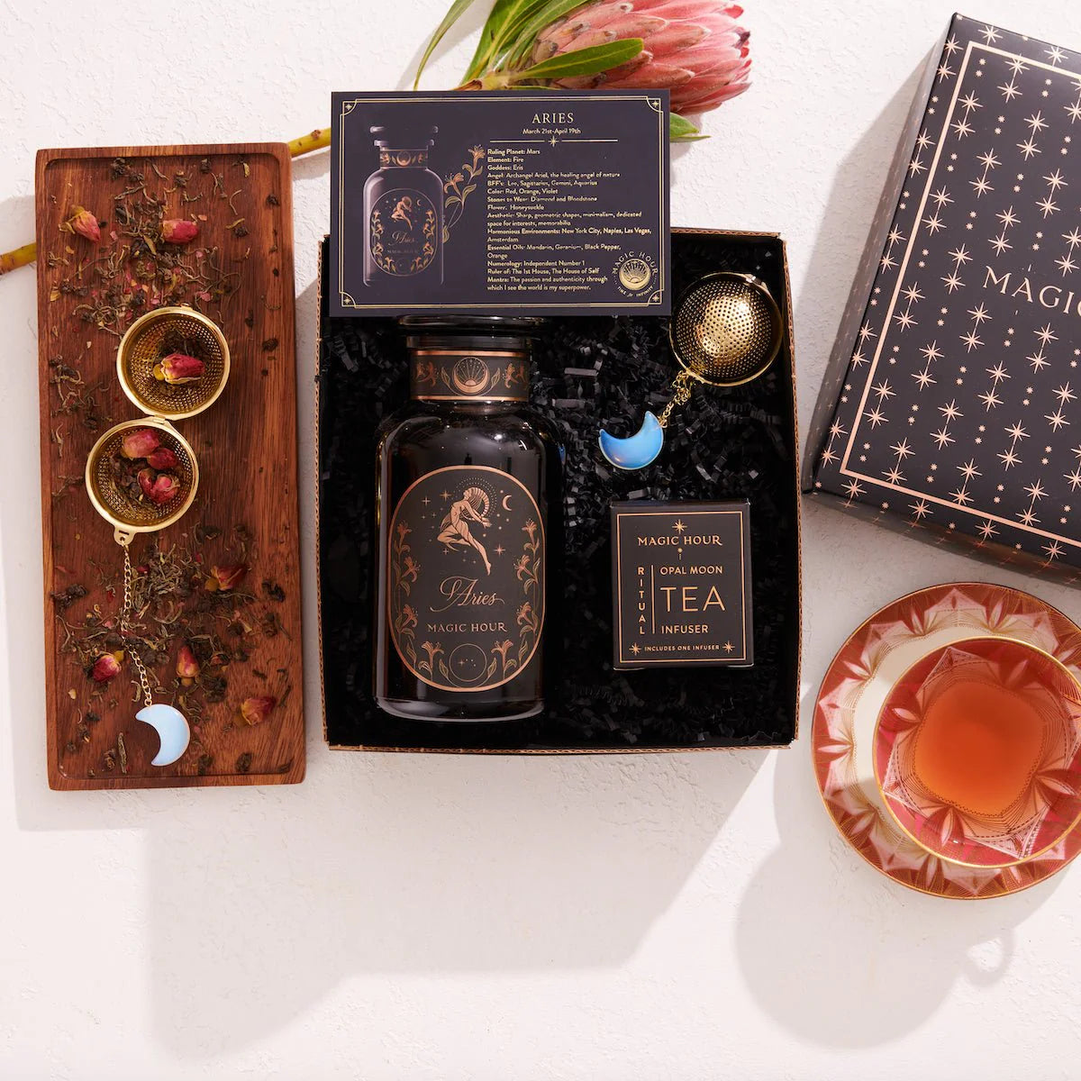 The Aries Astrology Tea Gift Set features a decorative tea jar, an Opalite Moon Tea Strainer, and a Magic Hour package accompanied by a card that reads "Aries." This set includes a wooden tray displaying tea leaves, rose petals, and blue crescent decorations. A steaming cup of tea elegantly sits on a saucer next to the delightful Aries Astrology Gift Set.