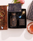 The Aries Astrology Tea Gift Set features a decorative tea jar, an Opalite Moon Tea Strainer, and a Magic Hour package accompanied by a card that reads "Aries." This set includes a wooden tray displaying tea leaves, rose petals, and blue crescent decorations. A steaming cup of tea elegantly sits on a saucer next to the delightful Aries Astrology Gift Set.