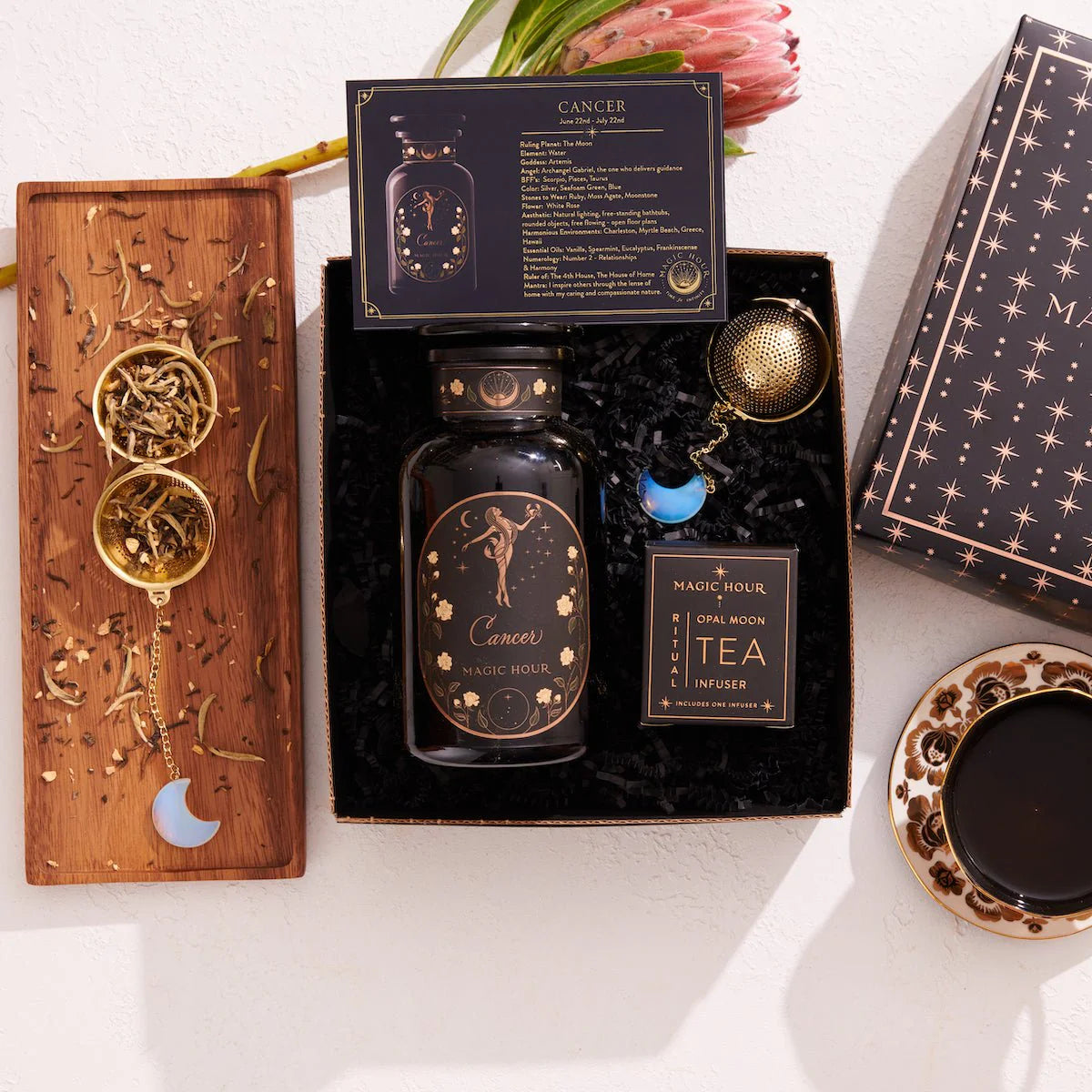 Explore the Cancer Astrology Tea Gift Set, a relaxing tea experience with a black tea jar designed with "Cancer" zodiac details. This charming set features an infuser and a "Full Moon Tea Infuser" box, two gold spoons with dried organic ingredients, all beautifully arranged on a wooden board.