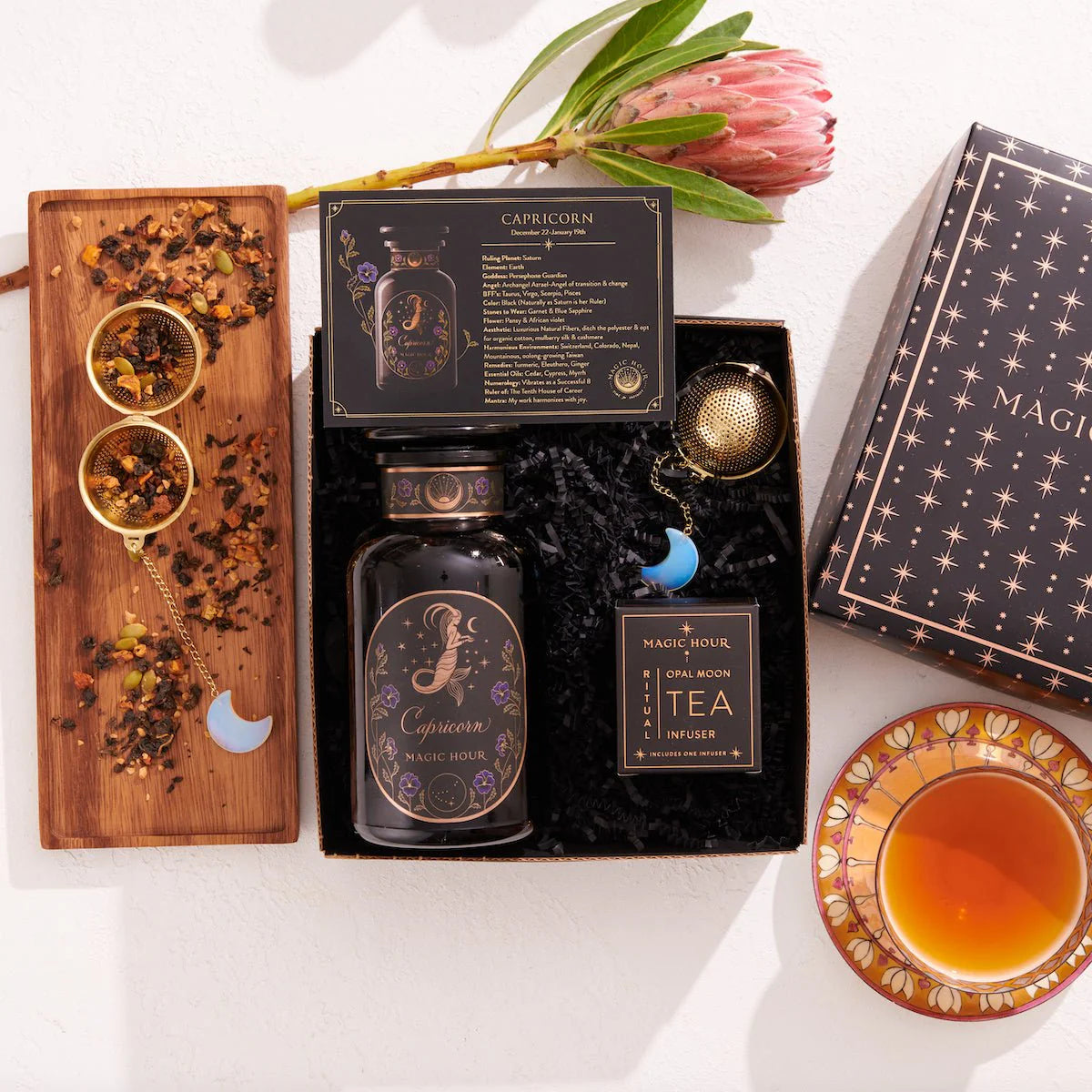 The Capricorn Astrology Tea Gift Set includes a tea canister, an Opalite Moon Tea Strainer, and packaging adorned with starry designs. A wooden tray artfully displays loose tea and scoops beside a steaming cup, with a single protea flower adding charm to this astrology-themed gift.