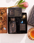 The Capricorn Astrology Tea Gift Set includes a tea canister, an Opalite Moon Tea Strainer, and packaging adorned with starry designs. A wooden tray artfully displays loose tea and scoops beside a steaming cup, with a single protea flower adding charm to this astrology-themed gift.