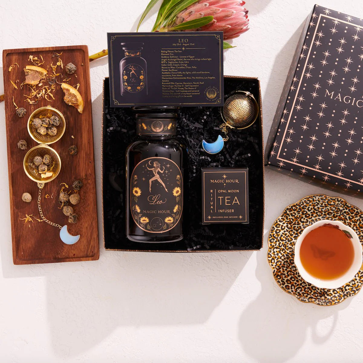 Presenting the Leo Astrology Tea Gift Set: a lively tea blend encased in a dark blue box featuring celestial patterns. This set includes a coordinating tin, an Opalite Moon Tea Strainer, dried herbs, and a teacup filled with tea, all beautifully highlighted by the elegant presence of a pink protea flower in the background.