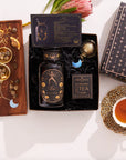 Presenting the Leo Astrology Tea Gift Set: a lively tea blend encased in a dark blue box featuring celestial patterns. This set includes a coordinating tin, an Opalite Moon Tea Strainer, dried herbs, and a teacup filled with tea, all beautifully highlighted by the elegant presence of a pink protea flower in the background.