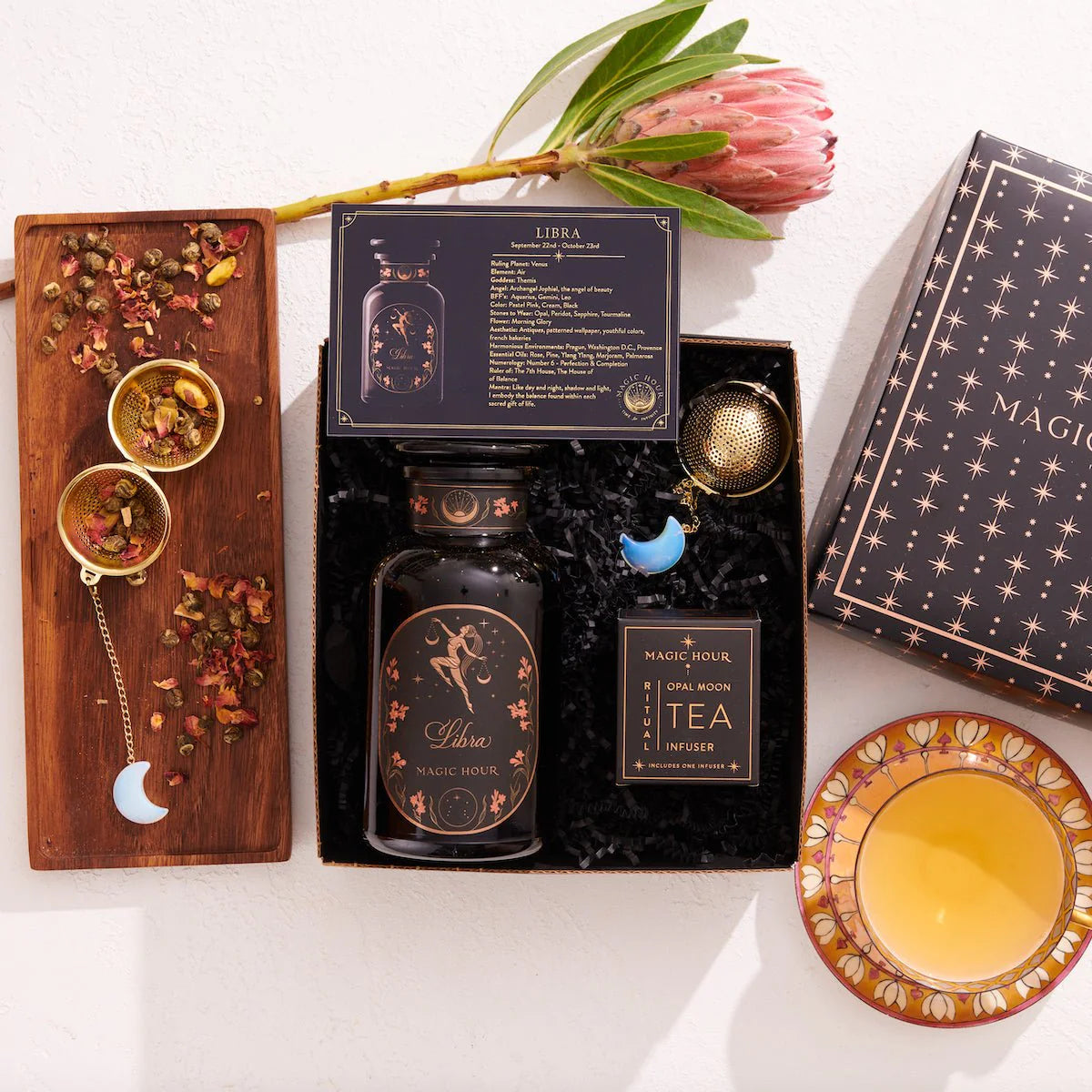 A flat lay showcasing the Libra Astrology Tea Gift Set. This set contains a dark tea jar adorned with floral designs, a box of Moonlight White Tea bags, a large decorative flower, two golden tea infusers, and a cup of tea on a patterned saucer. It also includes a booklet and packaging box to complete the ensemble.