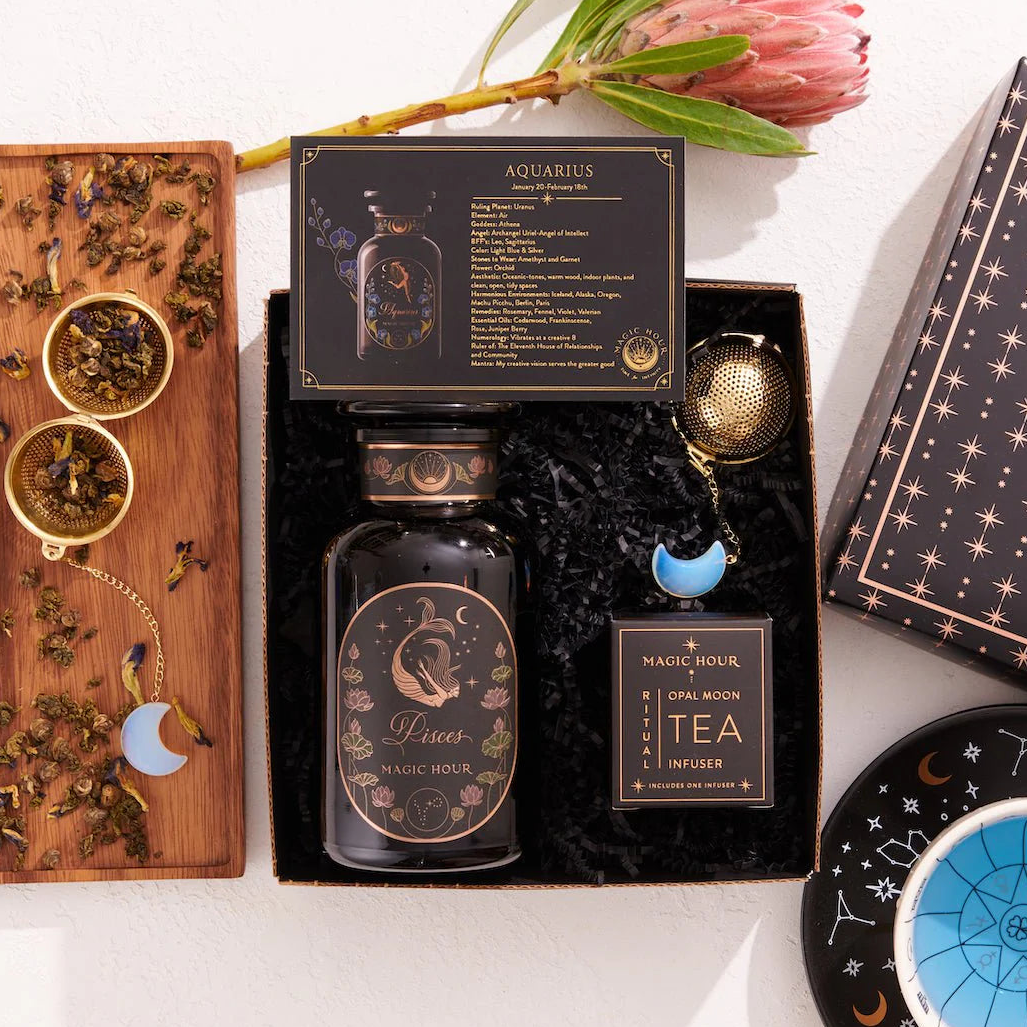 The Pisces Astrology Tea Gift Set includes a box of Aquarius tea, a bottle of Pisces tea, an Opalite Moon Tea Strainer, a blue celestial dish, and a wooden tray decorated with loose tea leaves and a protea flower. This captivating assortment is ideal for celestial enthusiasts.