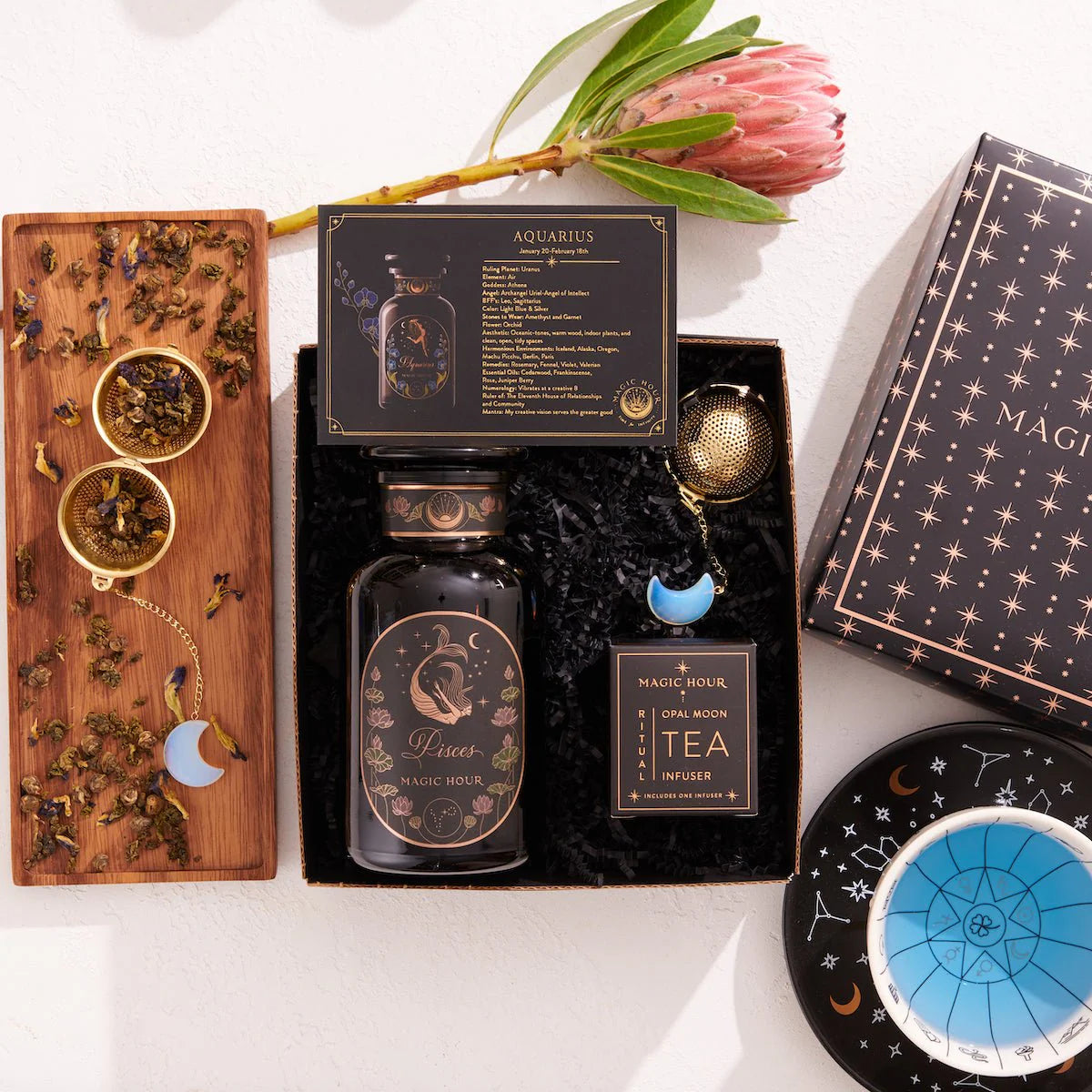 The Pisces Astrology Tea Gift Set includes a box of Aquarius tea, a bottle of Pisces tea, an Opalite Moon Tea Strainer, a blue celestial dish, and a wooden tray decorated with loose tea leaves and a protea flower. This captivating assortment is ideal for celestial enthusiasts.