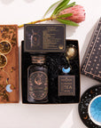 The Pisces Astrology Tea Gift Set includes a box of Aquarius tea, a bottle of Pisces tea, an Opalite Moon Tea Strainer, a blue celestial dish, and a wooden tray decorated with loose tea leaves and a protea flower. This captivating assortment is ideal for celestial enthusiasts.