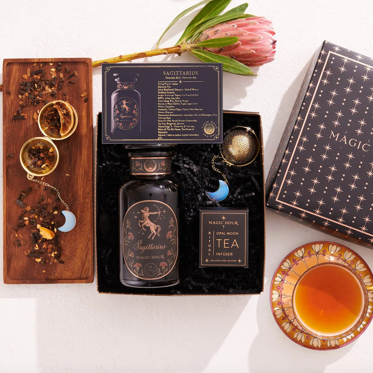 The Sagittarius Astrology Tea Gift Set is a decorative box featuring themed tea items such as a jar and an Opalite Moon Tea Strainer. Accompanied by a cup of tea on a wooden tray with dried herbs and a flower, the celestial packaging elevates this astrology tea experience.