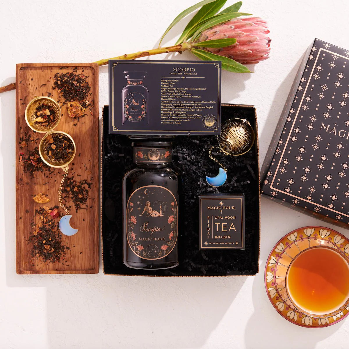 The Scorpio Astrology Tea Gift Set includes &quot;Scorpio&quot; tea, a wooden tray for loose leaf tea, and two gold tea infusers decorated with crescent moon charms. It is beautifully presented with a blooming protea flower and steaming cup of tea in a black box embellished with starry details.