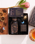 The Scorpio Astrology Tea Gift Set includes "Scorpio" tea, a wooden tray for loose leaf tea, and two gold tea infusers decorated with crescent moon charms. It is beautifully presented with a blooming protea flower and steaming cup of tea in a black box embellished with starry details.