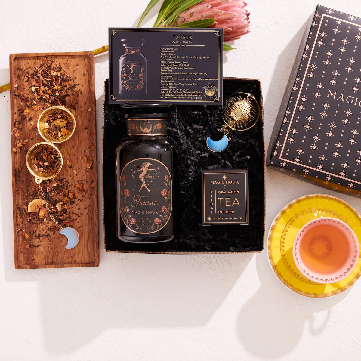 The Taurus Astrology Tea Gift Set offers a luxurious collection featuring a Taurus-themed tea blend, an elegant tea infuser, and precise measuring spoons displayed on a wooden tray decorated with loose tea leaves. Completing this celestial arrangement is a bright yellow teacup and saucer, alongside a black box adorned with star patterns.