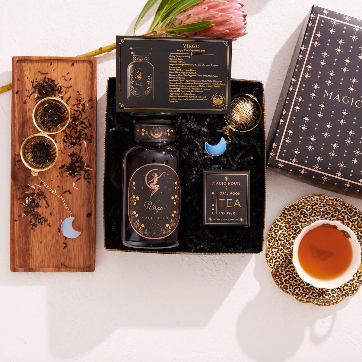 In a flat lay image of the Virgo Astrology Tea Gift Set, you can see a Virgo-themed bottle, two moon-shaped tea infusers, a box of Magic Hour full moon tea with organic ingredients, and a teacup with steeped tea, each item meticulously arranged on a white surface.