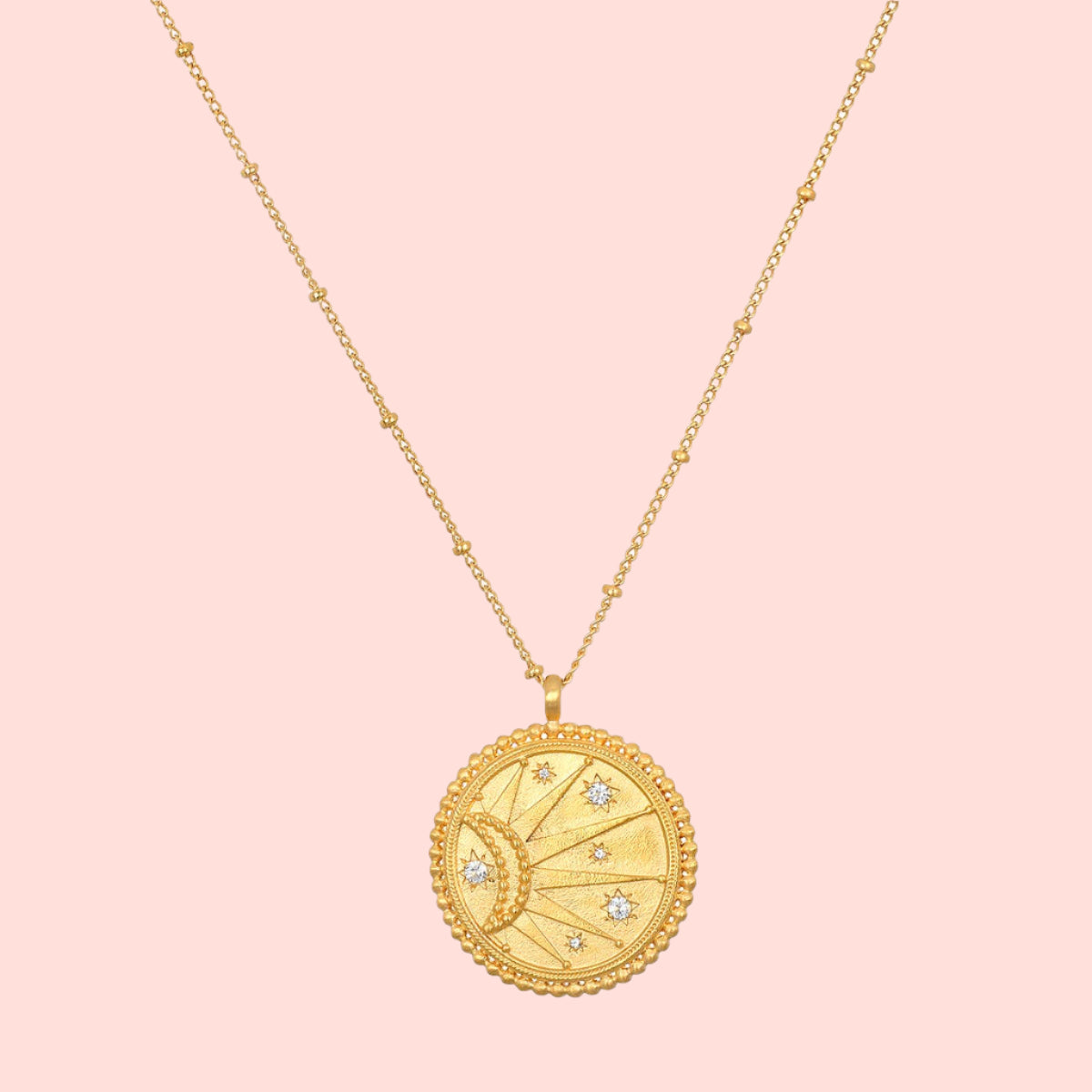 The 18" Sunburst Medallion Necklace features a gold plated round, textured pendant engraved with a crescent moon and white star accents, complemented by delicate bead details along the chain.