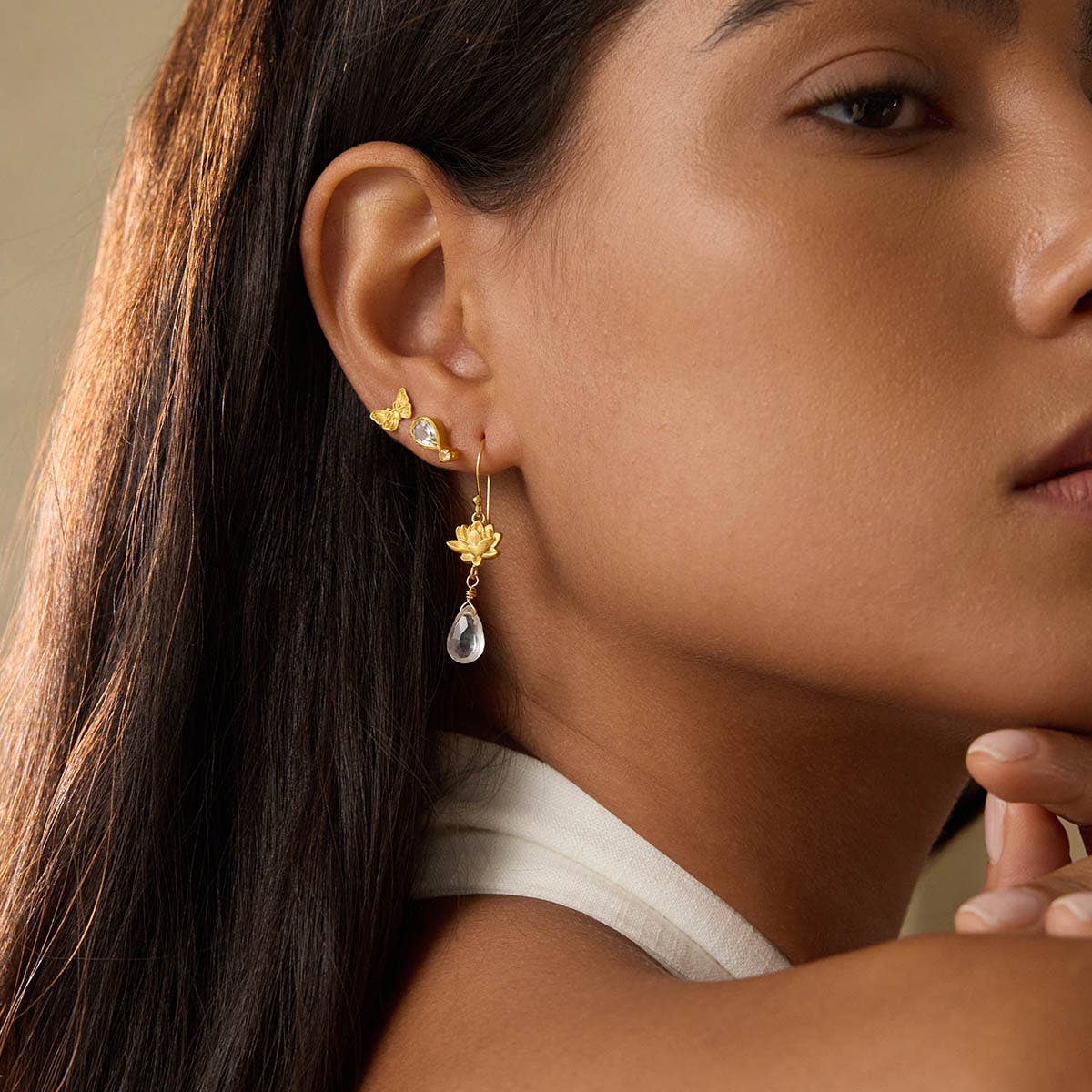 A woman with long brown hair, dressed in a sleeveless white top, wears 18k gold plated Spiritual Journey Lotus Crystal Earrings featuring a clear teardrop gem and two small crystal leaf-shaped accents.