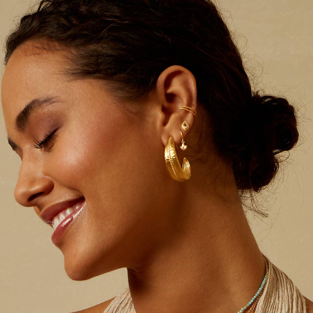 A woman with dark hair in a low bun smiles with her eyes closed, wearing multiple gold earrings—including a large textured hoop, Inner Light Blue Topaz Pear Stud Earrings, and smaller studs and cuffs. She wears a light-colored top.