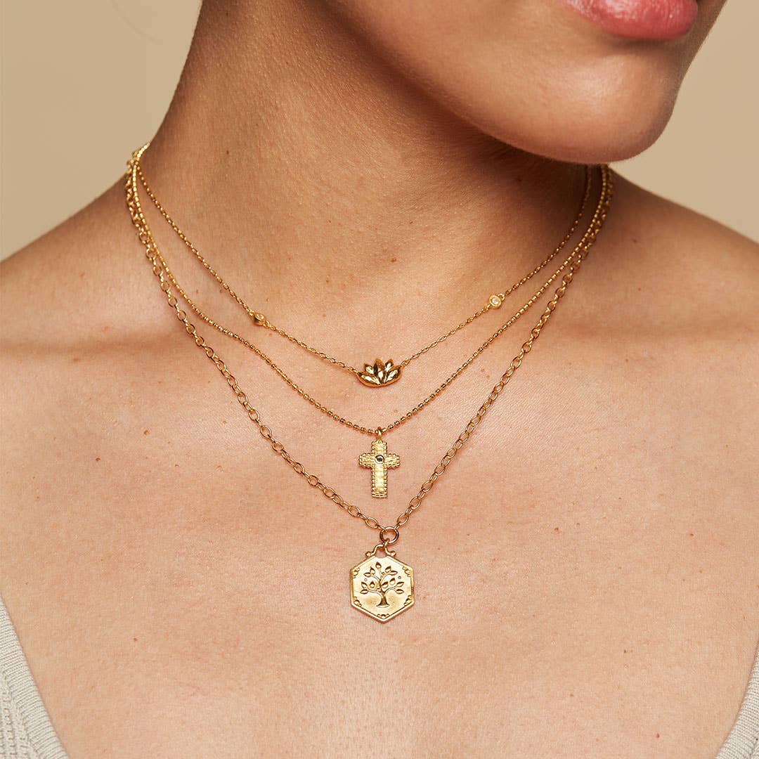 A woman wears a 16" Emerald Dot Cross Pendant Necklace with a 3" extension, styled with other gold layers and a light-colored top, highlighting her elegant faith-inspired jewelry look.