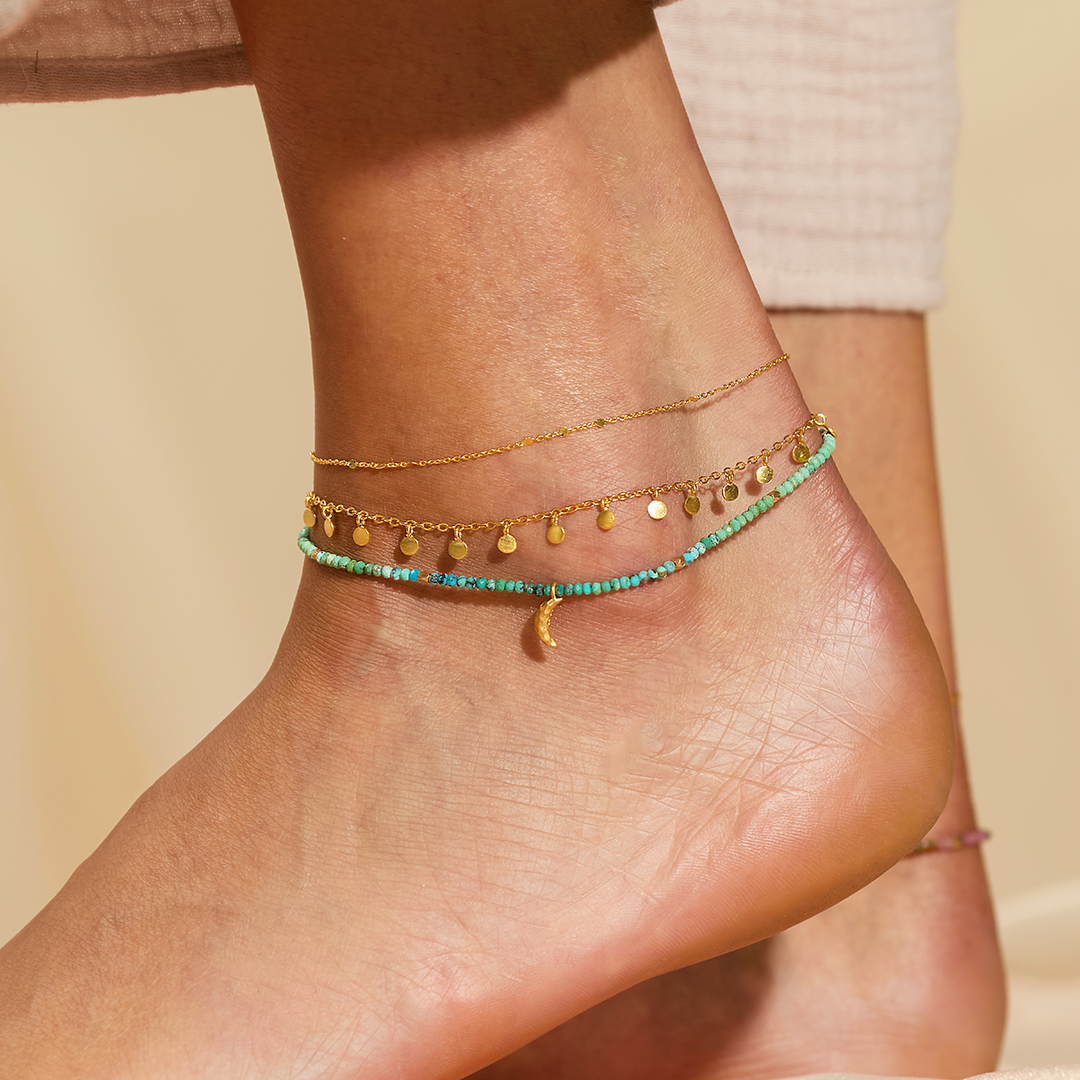 A close-up shows a person's ankle wearing three anklets: a delicate gold chain, a gold chain with small charms, and the 9" Turquoise moon anklet with beads, a small gold charm, and a 1" extender. The background is soft and neutral.