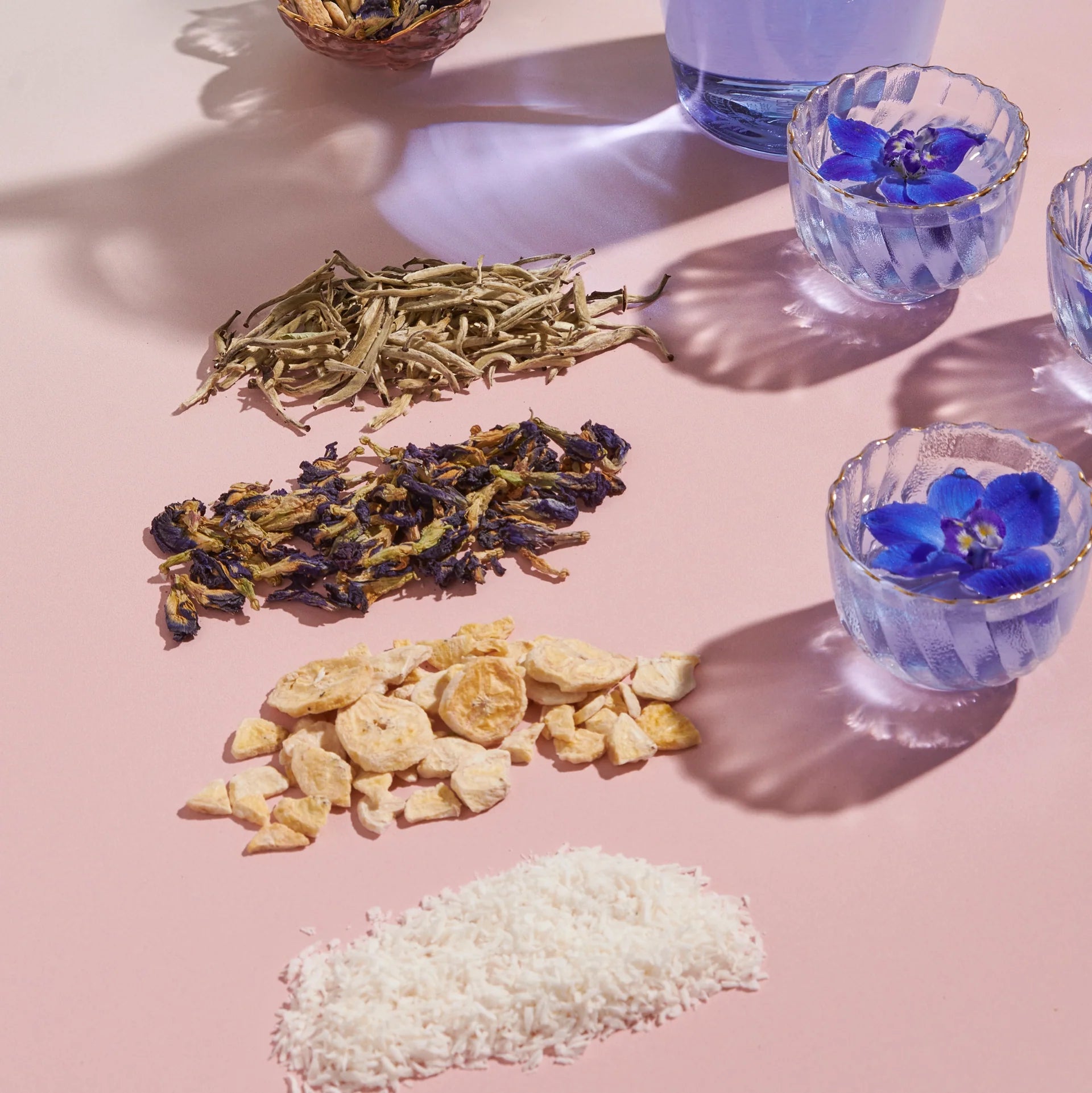 On a pink surface, four rows of dried ingredients from the Butterfly Blue Coconut Cream Iced White Tea Refill Pouch are displayed: white tea leaves, butterfly pea flowers, dried ginger pieces, and shredded coconut. Nearby, three glasses of iced tea show blue liquid with butterfly pea flowers. Shadows are cast nearby.