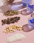 An arrangement of dried tea leaves, dehydrated fruit pieces, and shredded coconut displayed on a pink surface, with three glass cups of Magic Hour Butterfly Blue Coconut Cream Iced White Tea, one containing fresh butterfly pea flowers. Shadows cast over the scene from lighting create a refreshing summer hydration tableau.