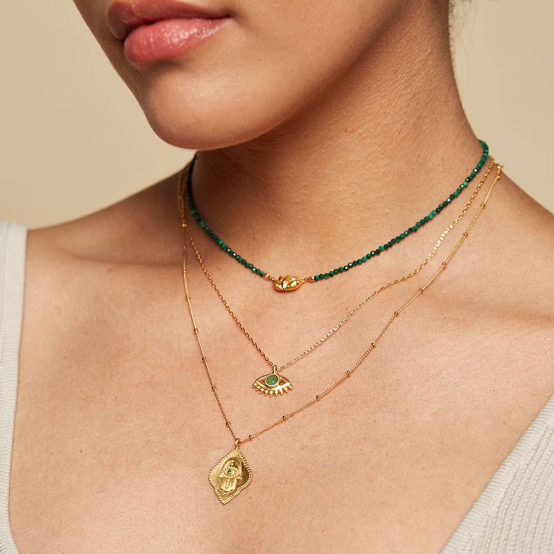 A close-up of a woman’s neck shows her layering necklaces, including the 16" Emerald Hamsa Pendant Necklace with a 3" extension chain. She pairs it with a green beaded choker, an evil eye gold chain, and wears a light top.