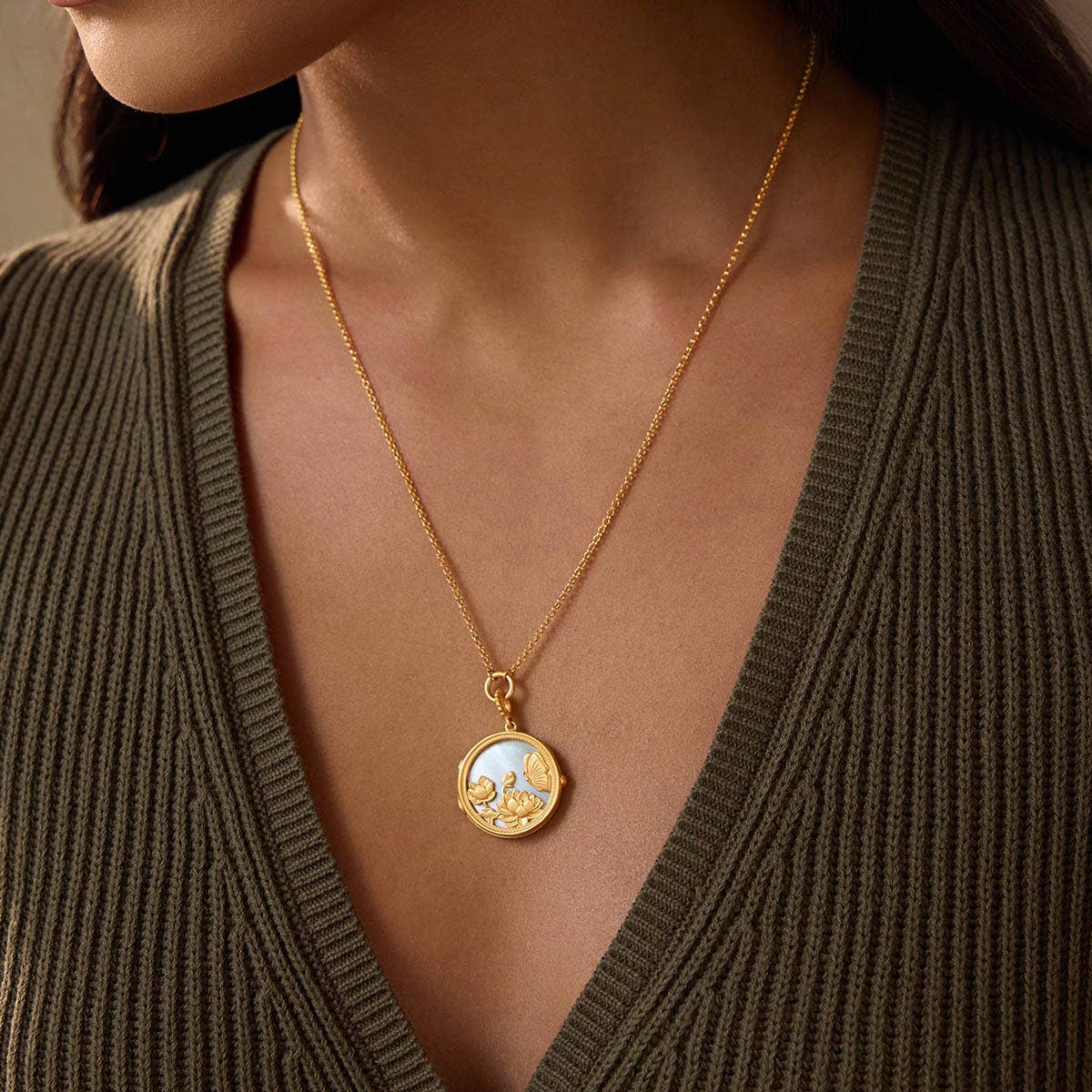 A woman wears a ribbed, olive green V-neck sweater and the Take Flight Butterfly Pearl Locket Necklace 18”, an 18k gold plated piece. The focus is on the necklace and upper chest, with her face partially visible.