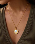 A woman wears a ribbed, olive green V-neck sweater and the Take Flight Butterfly Pearl Locket Necklace 18”, an 18k gold plated piece. The focus is on the necklace and upper chest, with her face partially visible.