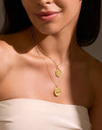 A woman wears a cream strapless top and two gold pendant necklaces—one being the Abundant Grounding Tree Necklace with a hexagonal charm, the other with a round coin-like charm—as she stands against a neutral background.