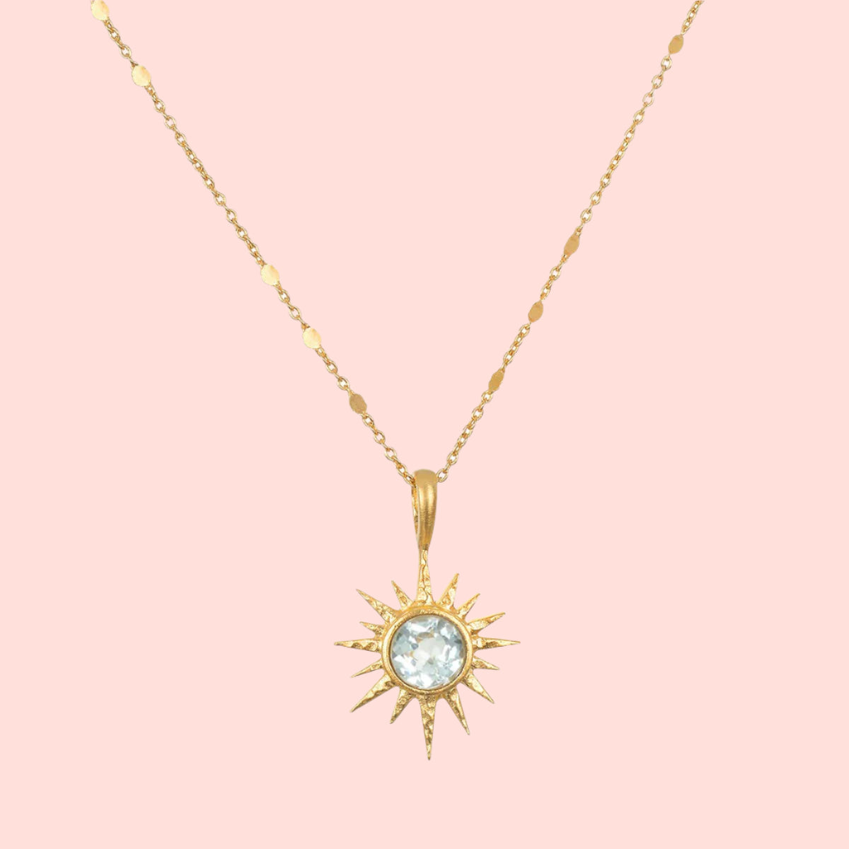 The 16" Blue Topaz North Star Necklace features a delicate 18k gold plated chain and a sun-shaped pendant with pointed rays, centered by a round, light blue topaz gemstone.