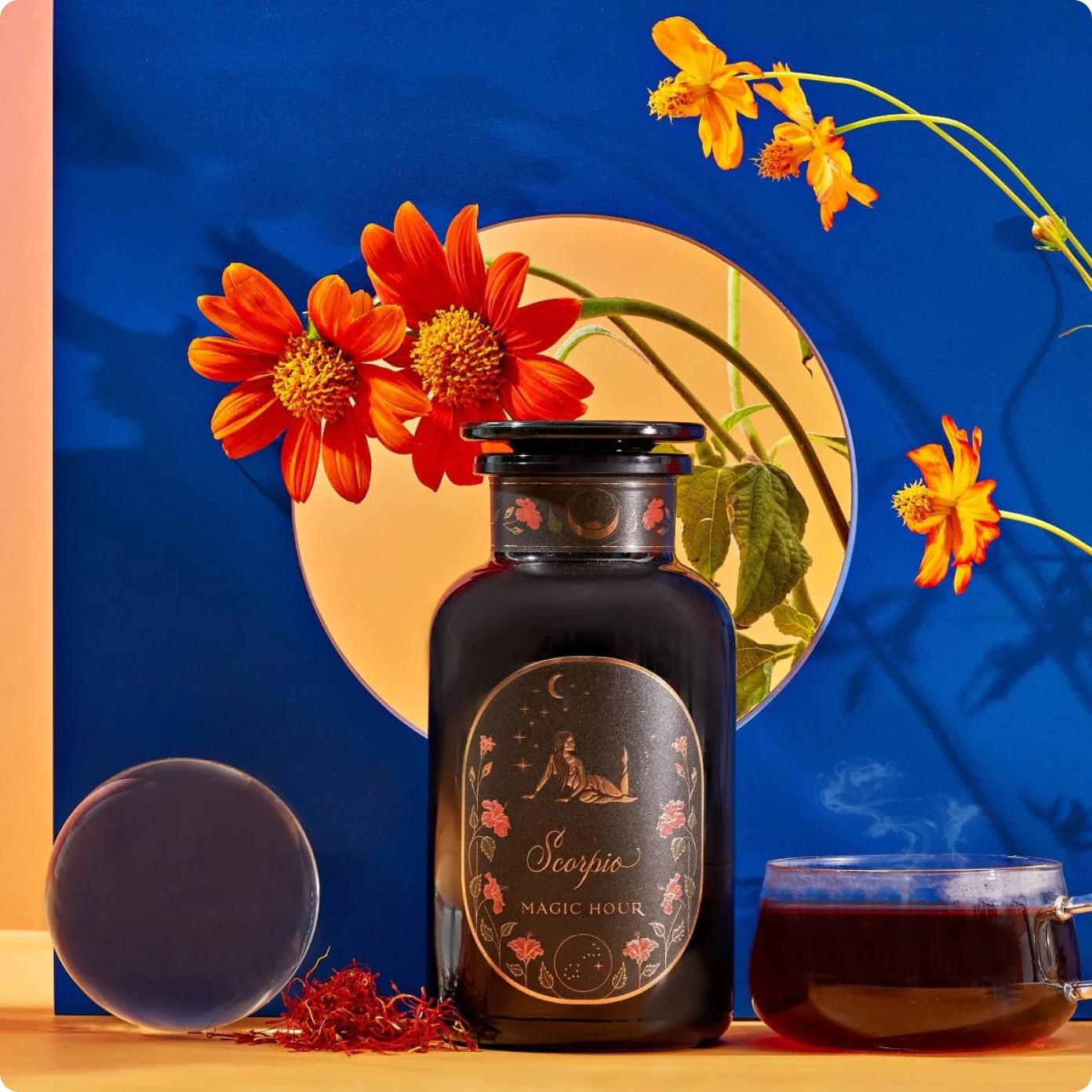 A dark glass jar labeled "Scorpio Magic Hour" is set in front of a blue and orange background with bright orange flowers. Beside it, a small clear cup contains a dark liquid. A few saffron threads and a circular glass object are also present in the scene.