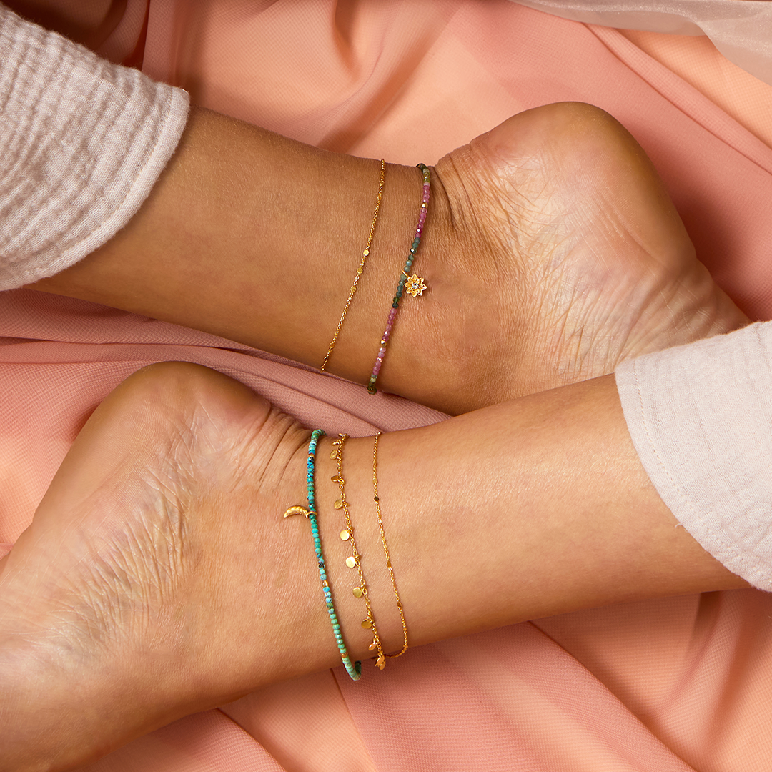 Two ankles, each wearing a 9" shimmering chain anklet with a 1" extender, rest on soft peach fabric. The gold plated anklets feature turquoise, pink, and gold tones accented with star and crescent moon charms.