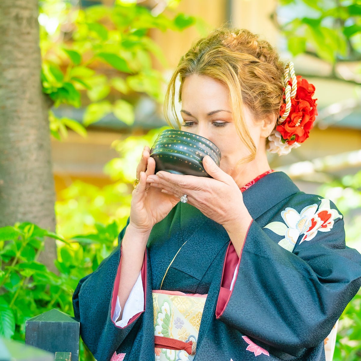 A woman dressed in a traditional Japanese kimono stands outside, surrounded by greenery. She is sipping from a patterned black bowl included in the Magic Hour Starter Kit by Magic Hour, holding it with both hands as part of a tea ceremony. Her kimono is dark with floral patterns, and she has decorative red and white flowers in her hair.