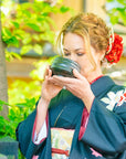 A woman dressed in a traditional Japanese kimono stands outside, surrounded by greenery. She is sipping from a patterned black bowl included in the Magic Hour Starter Kit by Magic Hour, holding it with both hands as part of a tea ceremony. Her kimono is dark with floral patterns, and she has decorative red and white flowers in her hair.