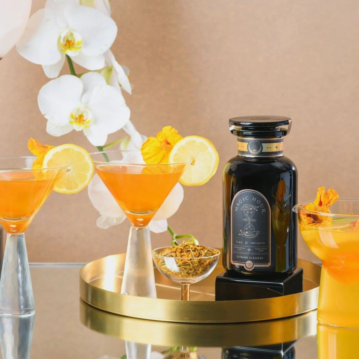 A bottle of Magic Hour tea concentrate is placed on a golden tray alongside two cocktails garnished with lemon slices and flowers. White orchids serve as a backdrop. An additional small dish with dried flowers is also on the tray. The setting is elegant and stylish.