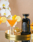 A bottle of Magic Hour tea concentrate is placed on a golden tray alongside two cocktails garnished with lemon slices and flowers. White orchids serve as a backdrop. An additional small dish with dried flowers is also on the tray. The setting is elegant and stylish.