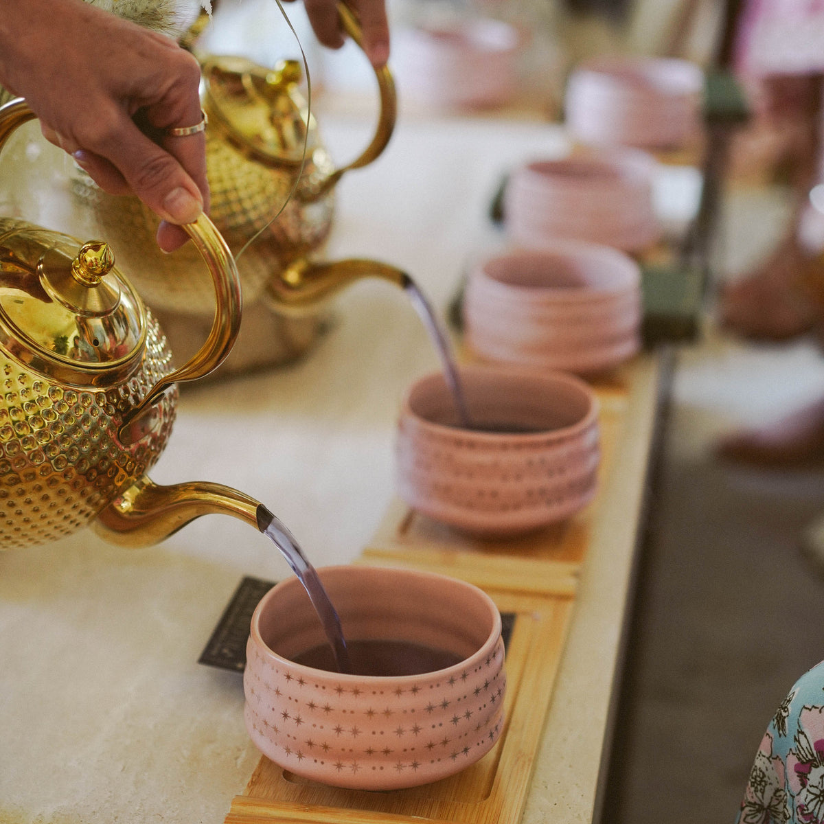 Captured in the soft glow of morning, a hand pours tea as part of The Soul of Tea: A Two Day Retreat in Ojai, CA, filling pink ceramic cups arranged neatly on a wooden tray—a moment that evokes the warmth and intimacy of an authentic tea ceremony.