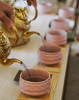 Captured in the soft glow of morning, a hand pours tea as part of The Soul of Tea: A Two Day Retreat in Ojai, CA, filling pink ceramic cups arranged neatly on a wooden tray—a moment that evokes the warmth and intimacy of an authentic tea ceremony.