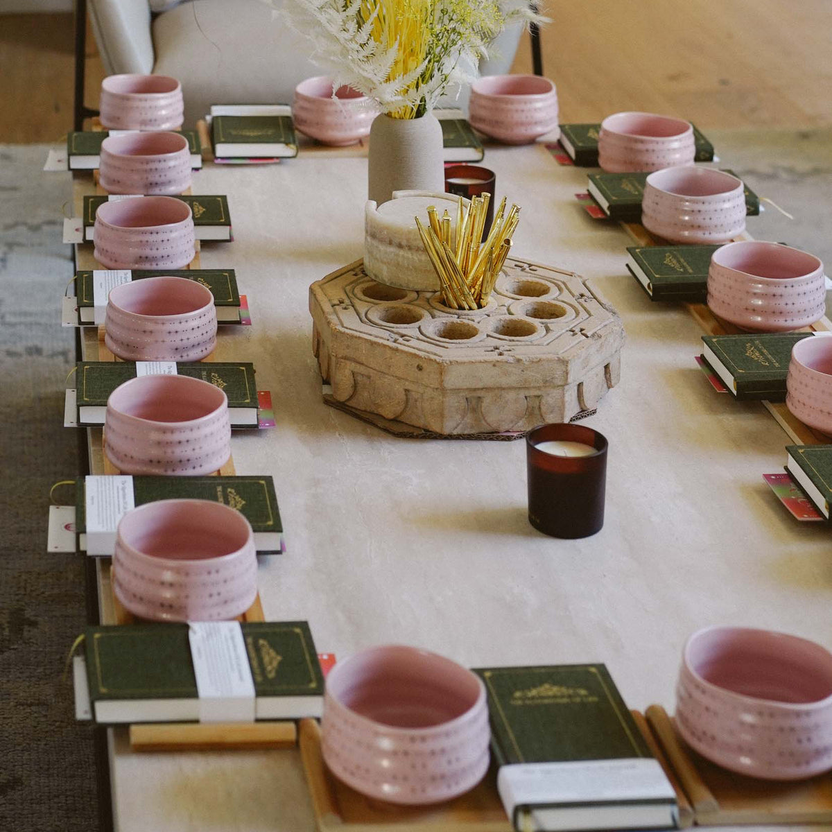 A rectangular table set with pink bowls, green books, candles, and a decorative centerpiece with yellow flowers is arranged neatly for a group gathering—perfect for The Soul of Tea: A Two Day Retreat in Ojai, CA.