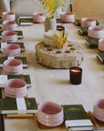 A rectangular table set with pink bowls, green books, candles, and a decorative centerpiece with yellow flowers is arranged neatly for a group gathering—perfect for The Soul of Tea: A Two Day Retreat in Ojai, CA.