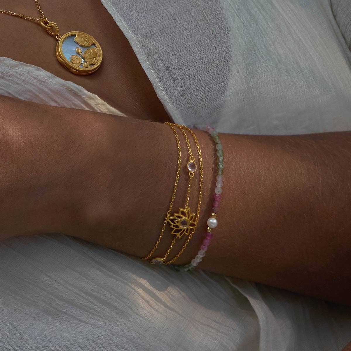 A person wears layered 18k gold plate bracelets, including the Transformed by Love Lotus Bracelet with a lotus flower charm and a pearl accent, plus a detailed gold pendant necklace, all against white textured fabric.